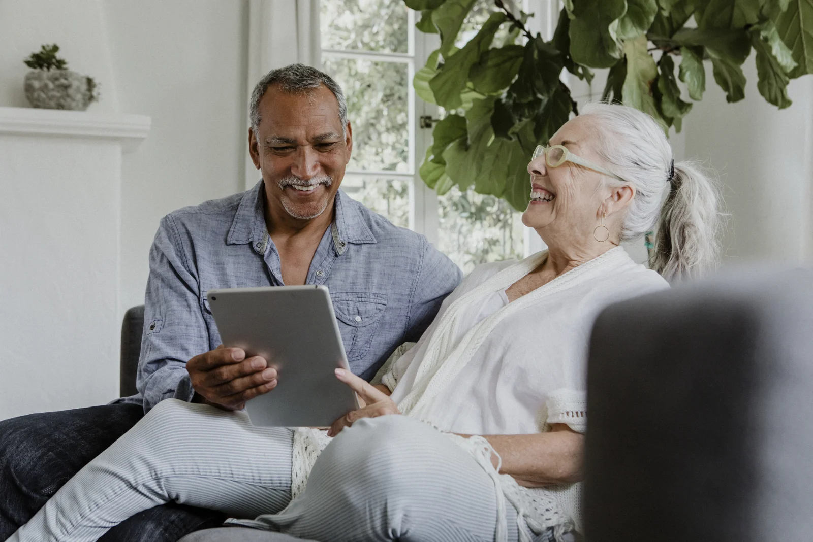 Casal de pessoas idosas sorrindo, usando um tablet juntos em ambiente claro e acolhedor